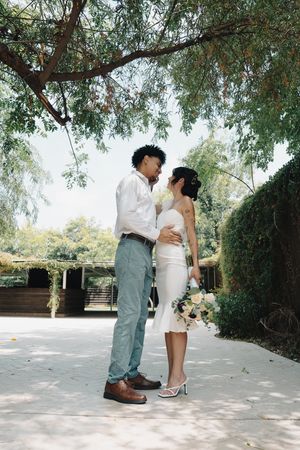 Interracial couple in wedding attire stands face to face under a canopy of green trees outdoors.