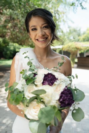 Beaming bride in white dress holds a stunning bouquet of white roses and purple hydrangeas outdoors.