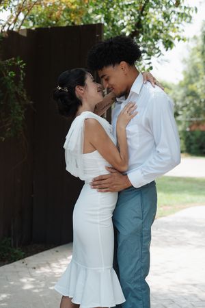 Interracial newlywed couple embraces closely, nearly kissing, in a lush outdoor garden setting.