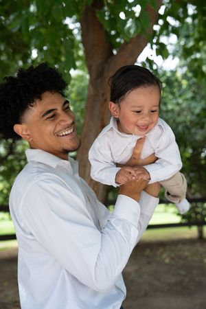 Laughing young father lifts his smiling baby boy outdoors under lush green trees.