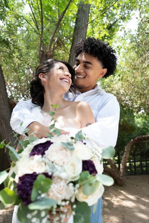 Joyful interracial newlyweds embrace under trees, bride holding a white and purple floral bouquet.