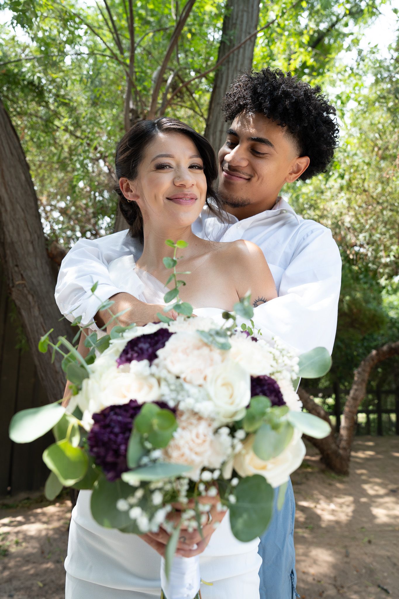 Happy interracial couple embraces outdoors, bride holding a bouquet of white and purple flowers.
