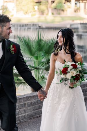 Bride and groom hold hands outdoors, bride in lace gown carrying red and white rose bouquet.