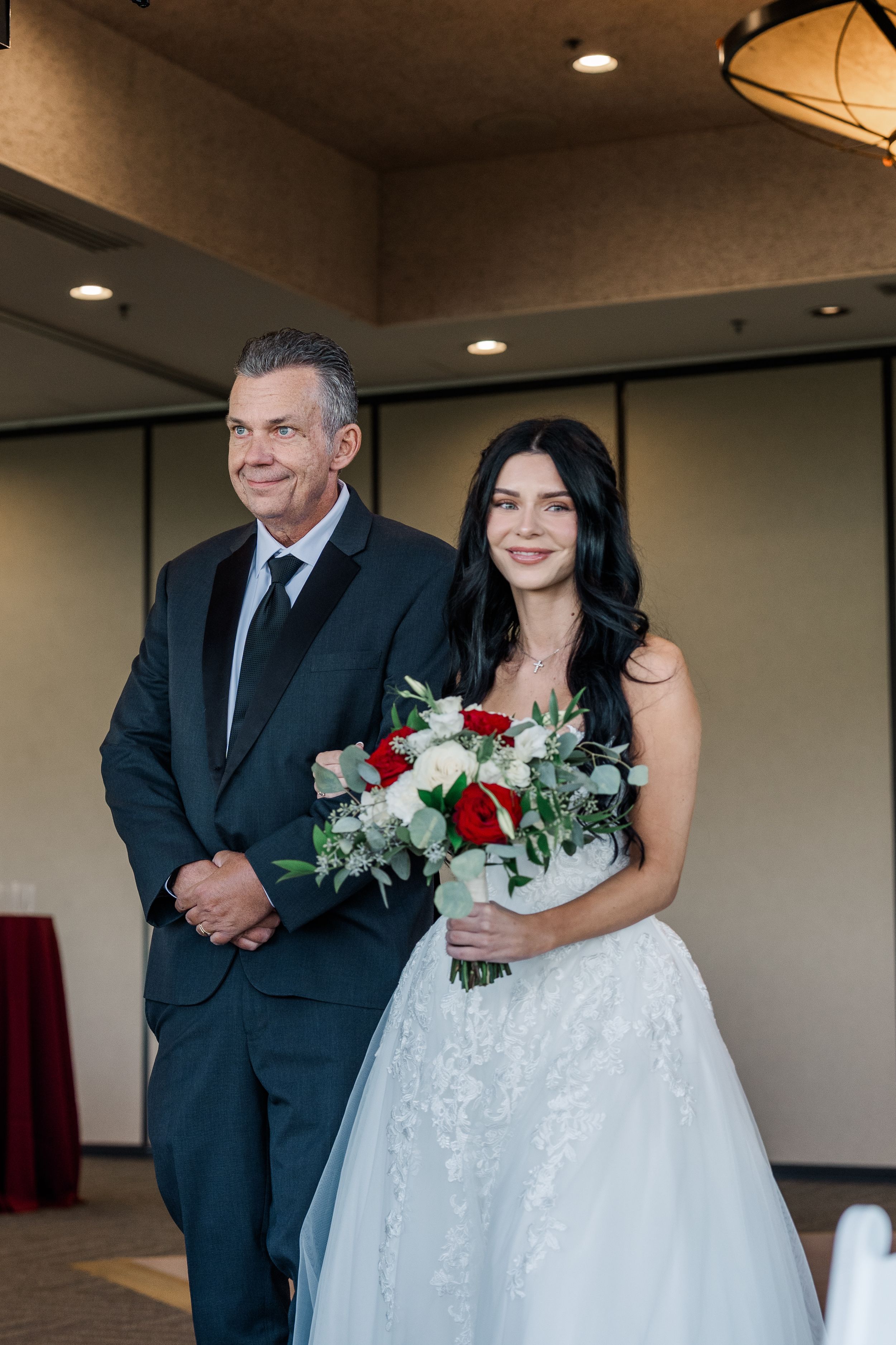 Bride in strapless lace gown holding red and white bouquet walks the aisle with her father.