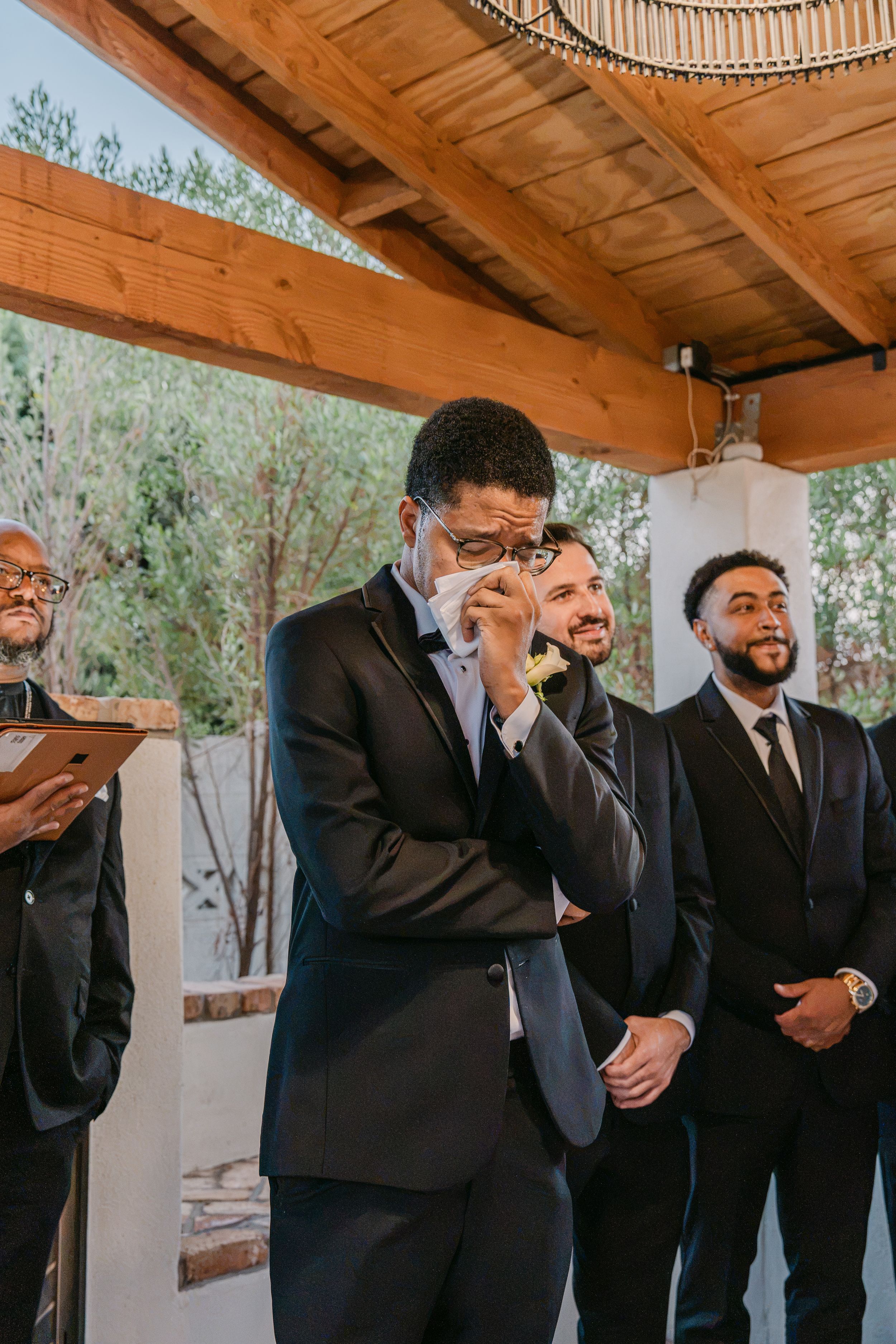Emotional groom wipes tears at altar surrounded by smiling groomsmen during outdoor wedding ceremony.