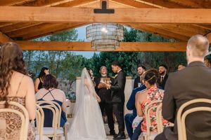 Bride and groom exchange vows under wooden pergola with chandelier before seated wedding guests.