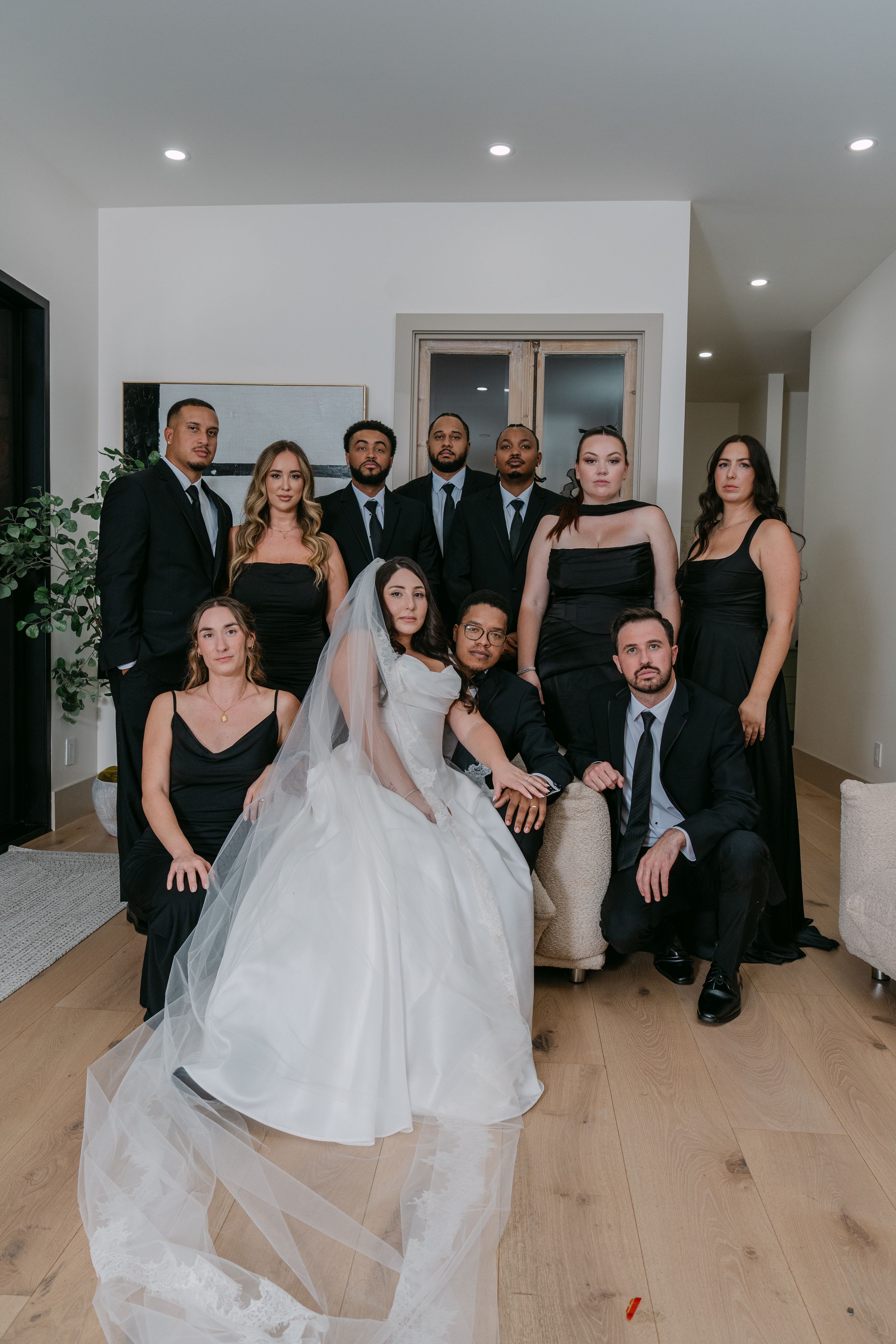 Wedding party in black attire poses with bride in white gown and veil in elegant indoor setting.