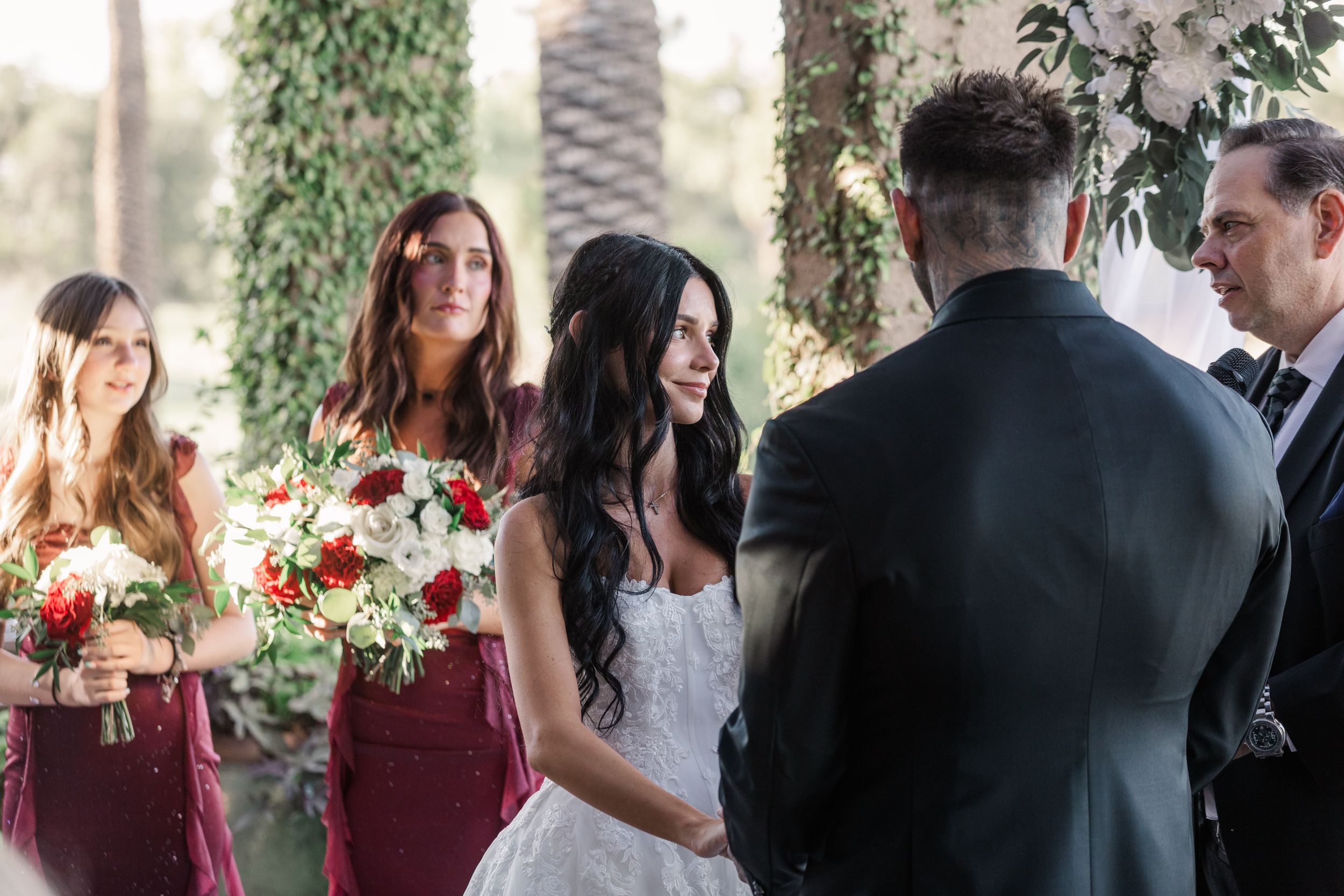 Bride in strapless lace gown smiles at groom during outdoor wedding ceremony with bridesmaids.