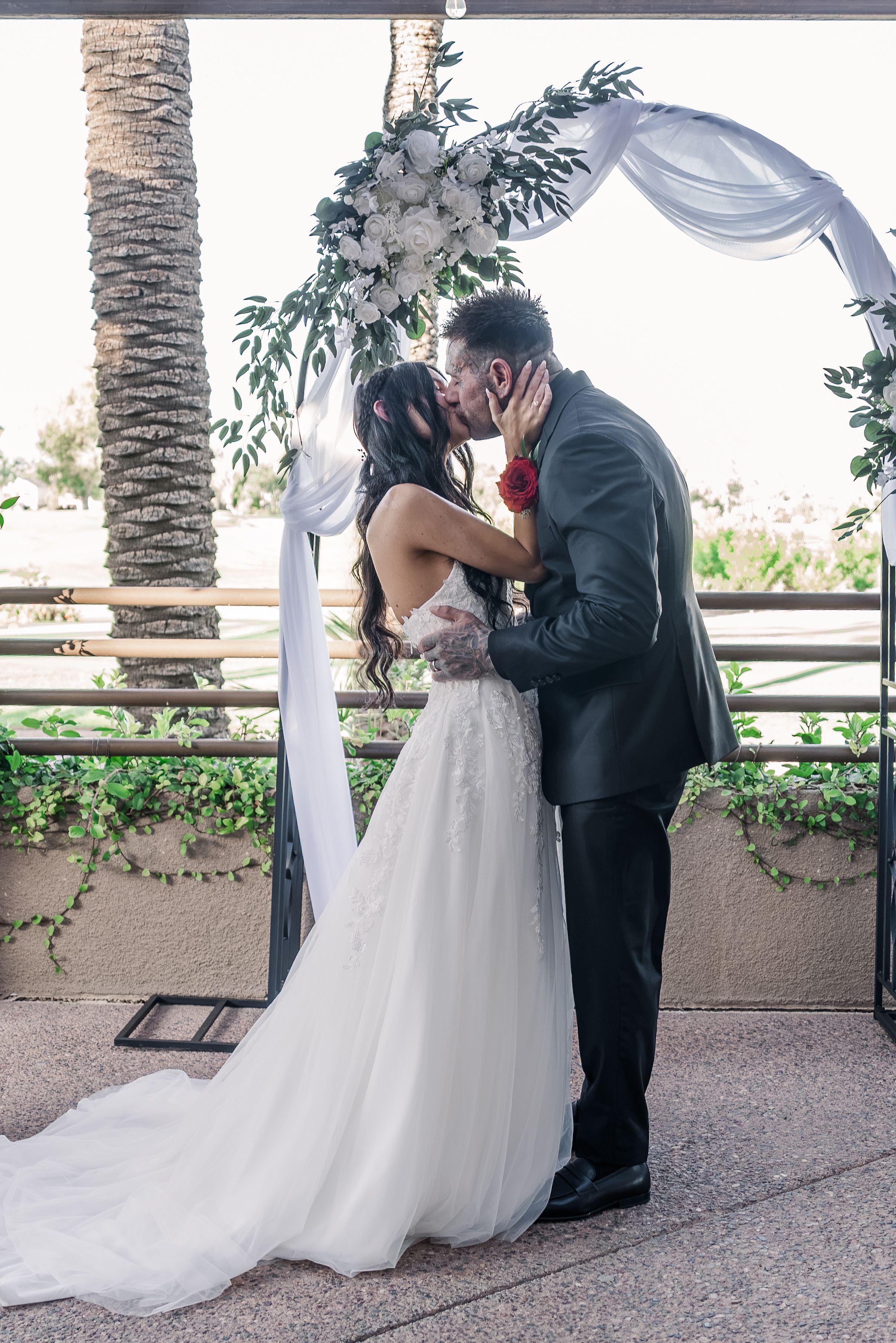 Bride and groom share first kiss under a floral arch adorned with white roses and greenery.
