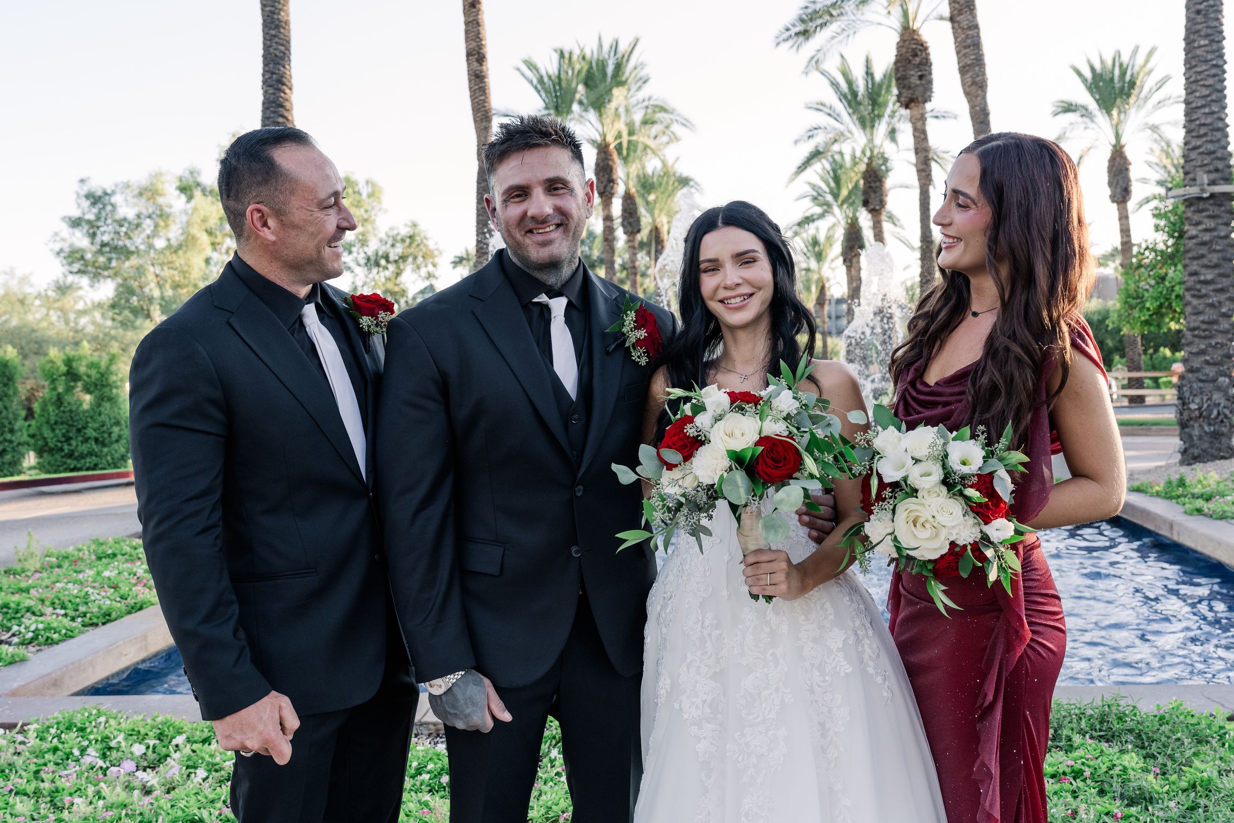 Wedding party of four smiling outdoors, bride in white gown holding red and white bouquet.