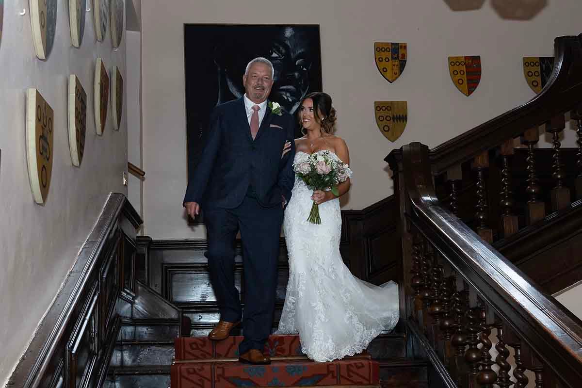 Bride in white lace gown and groom in blue suit descend ornate wooden staircase in elegant castle venue.