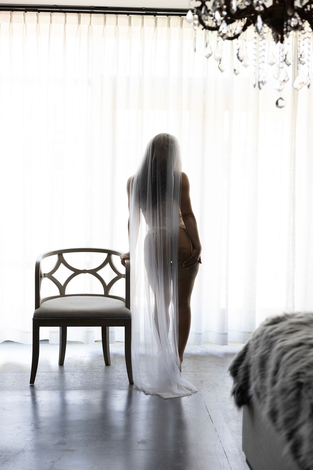 Silhouette of a bride wearing a long veil and lingerie standing before bright white curtains in an elegant room.