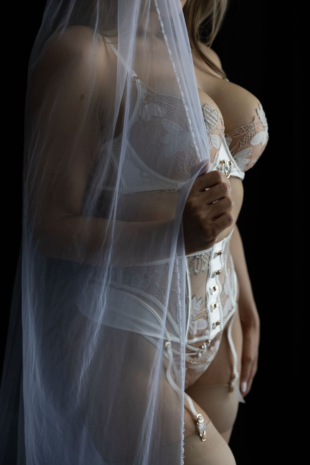 Woman in white bridal lingerie and corset holding a sheer veil against a dark background.