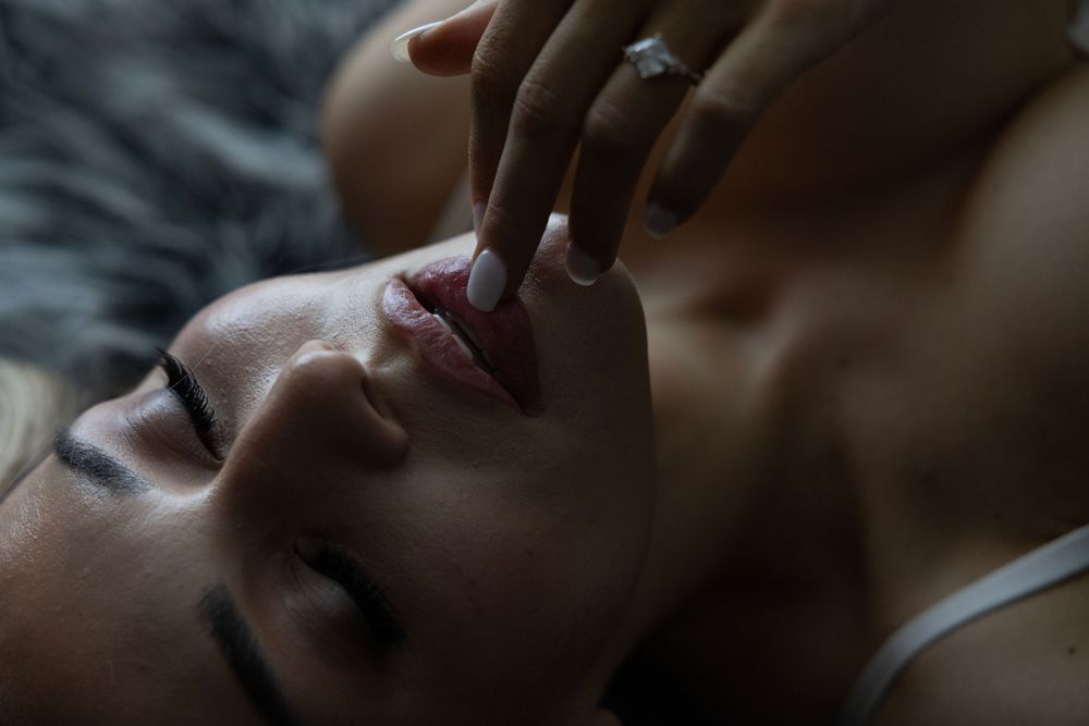 Close-up of a woman lying down with eyes closed as a hand gently touches her lips in soft lighting.
