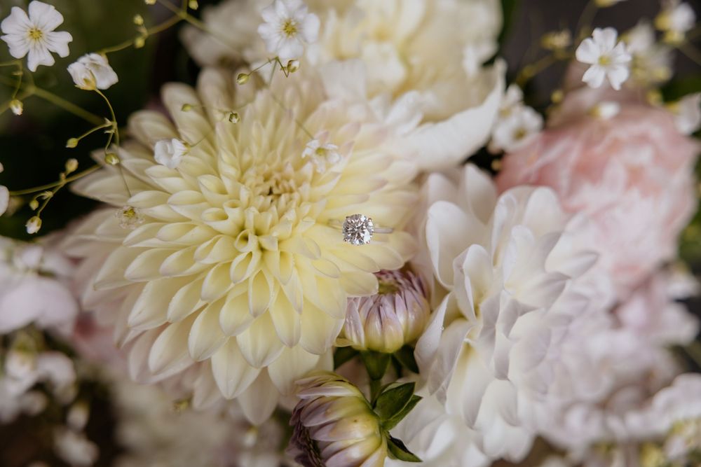Diamond solitaire engagement ring displayed among cream dahlias and delicate white baby's breath in bridal bouquet.