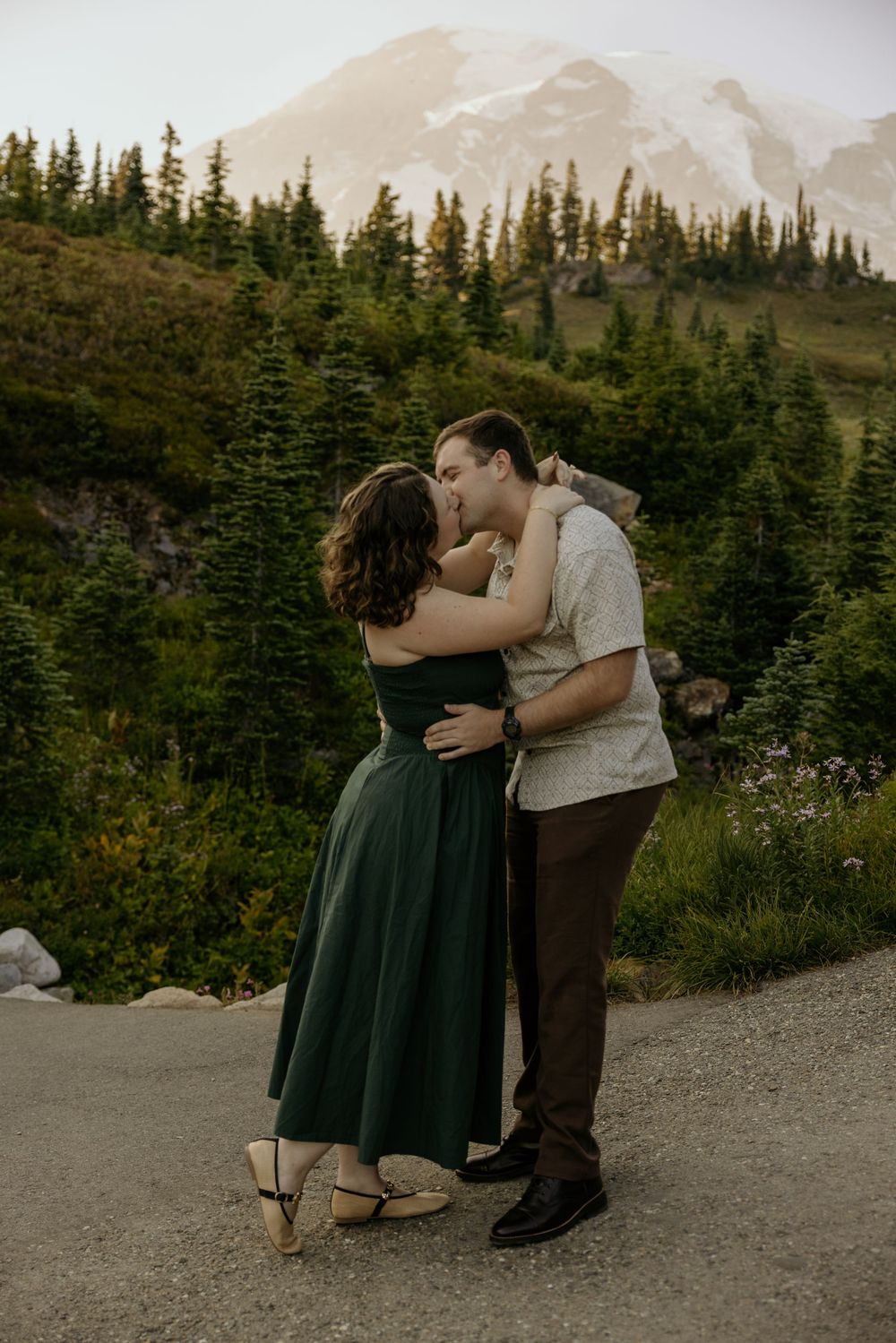 Couple sharing a romantic kiss at Mount Rainier, woman in green dress, surrounded by evergreen trees and snow-capped mountain.