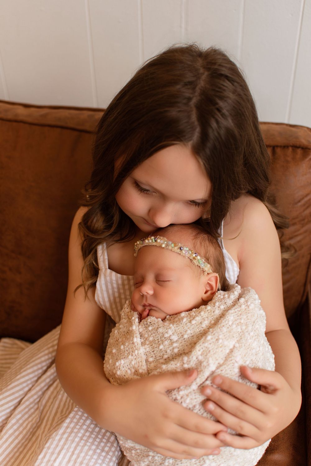 A young girl tenderly holds a sleeping newborn baby wrapped in a lace swaddle on a leather couch.