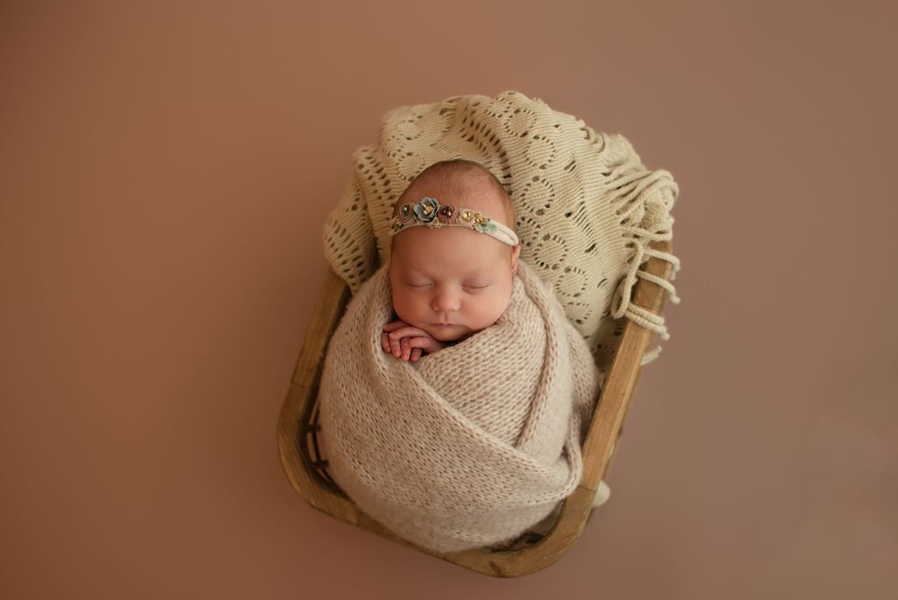 Newborn baby girl swaddled in beige wrap, sleeping peacefully in a wooden crate with knit blanket and floral headband.