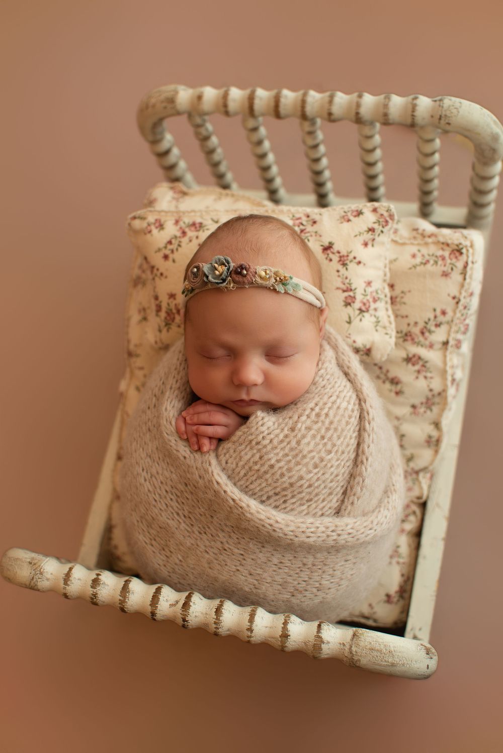 Sleeping newborn baby swaddled in beige knit wrap with floral headband lying in vintage white mini crib.