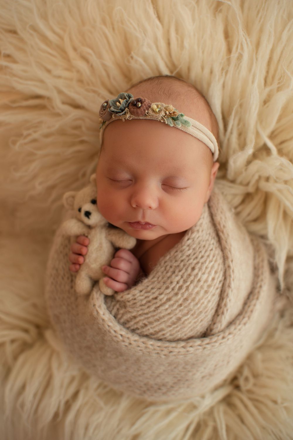 Newborn baby wrapped in beige knit swaddle holding tiny teddy bear with jeweled headband on fluffy rug.