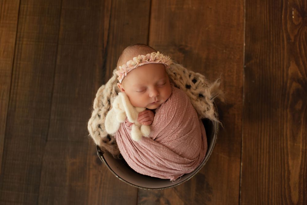 Newborn baby wrapped in pink swaddle sleeping in a rustic bucket with a floral headband on wood floor.