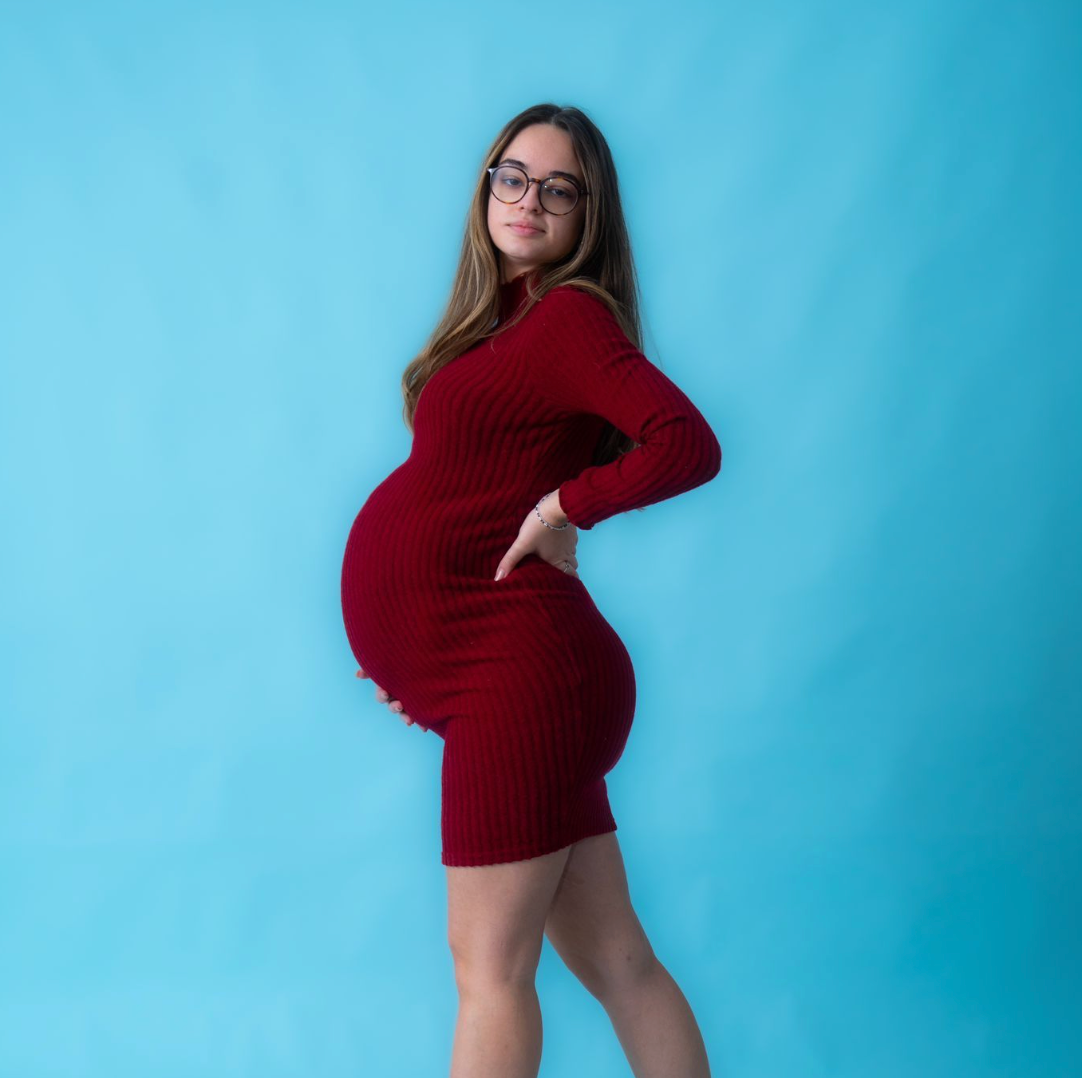 Pregnant woman in red ribbed dress posing against blue background, showcasing baby bump in maternity photo shoot.