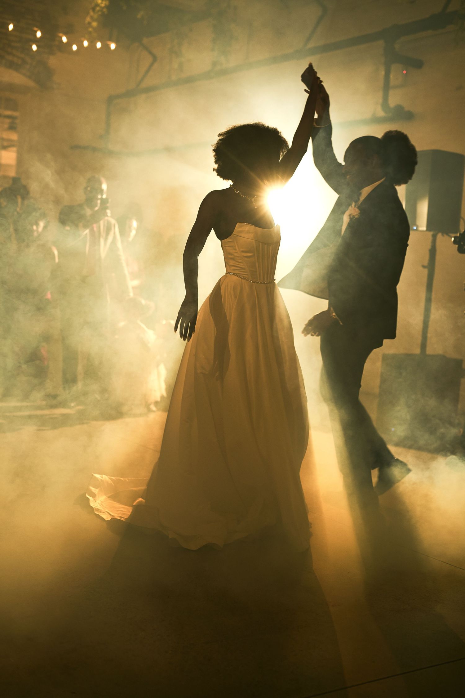 Silhouetted bride and groom sharing first dance in golden smoky light at romantic wedding reception.