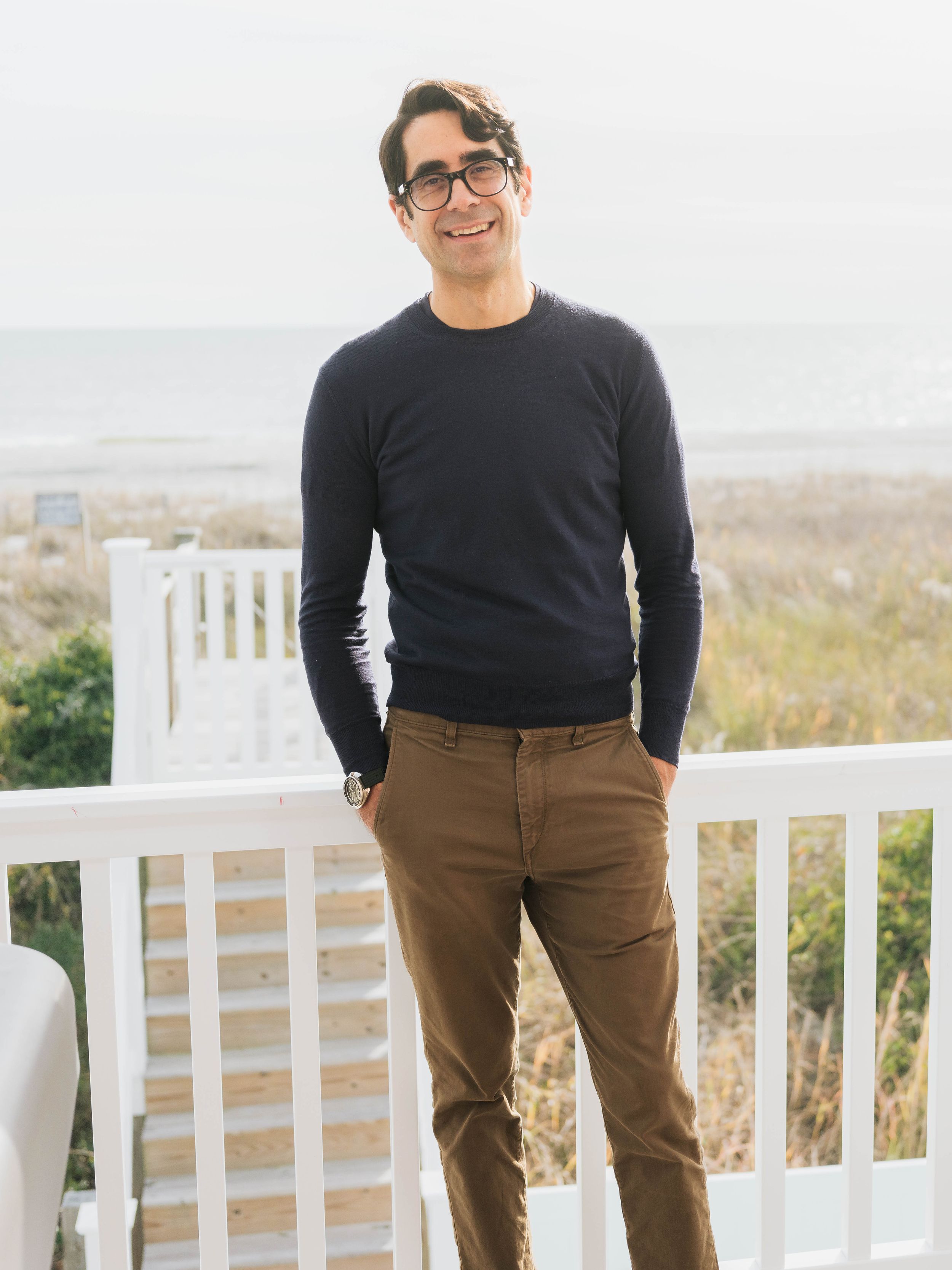 Man in navy sweater and brown chinos standing on a white railing deck with ocean view backdrop.