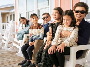 Multi-generational Asian family smiling together on a porch bench, including children and an elderly woman wearing sunglasses.