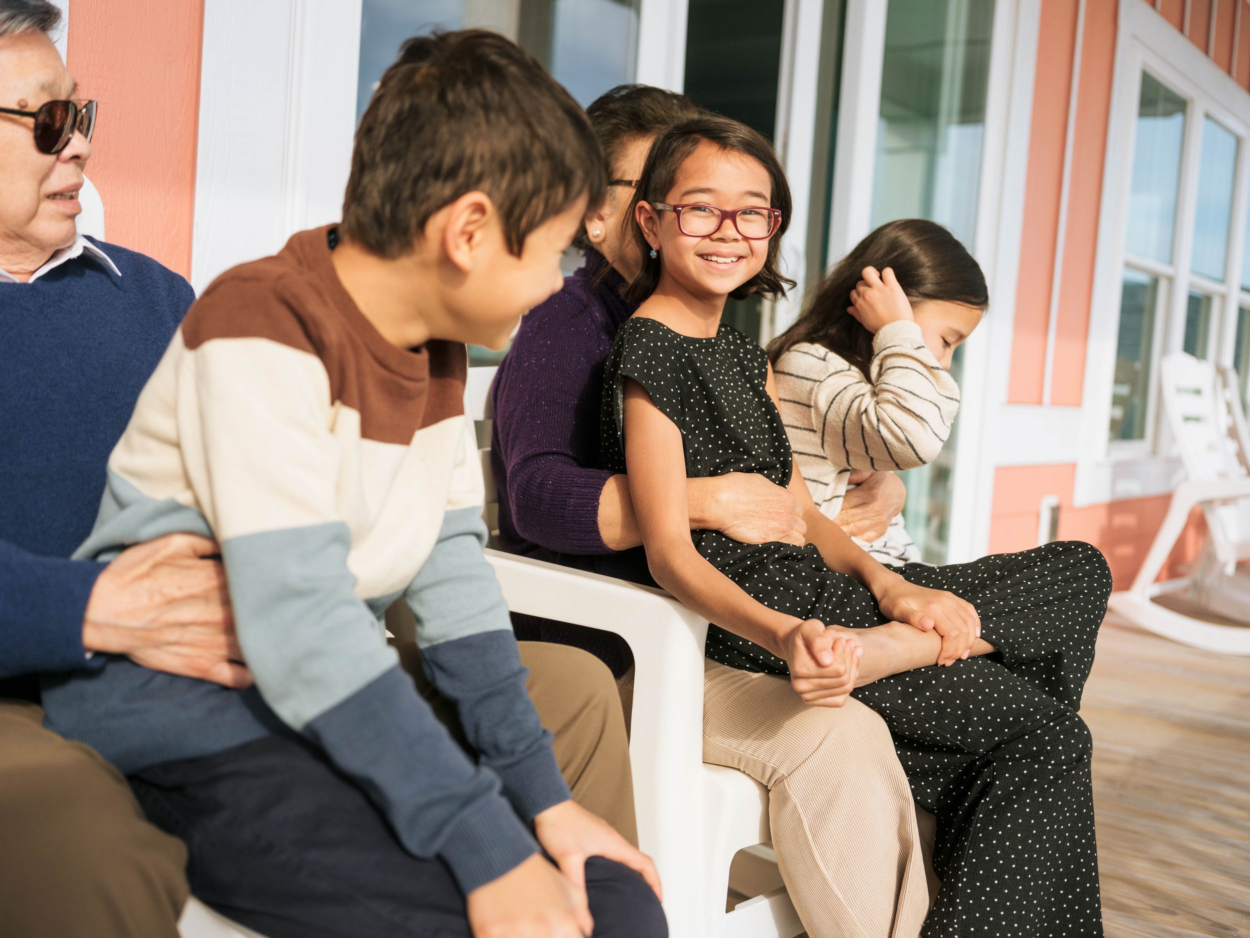 Asian family sitting together on a porch bench, smiling and enjoying quality time outdoors on a sunny day.