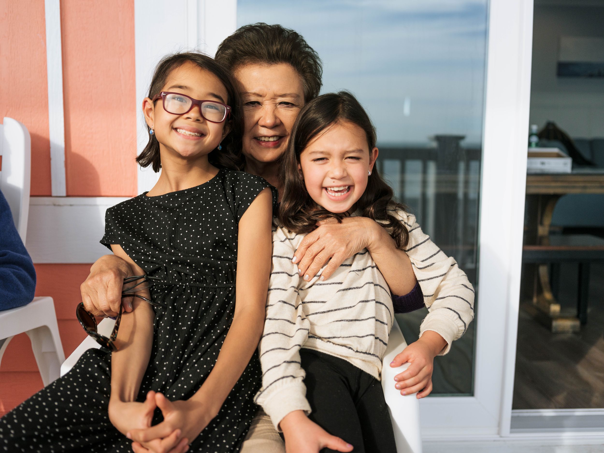 Grandmother smiling and hugging two young girls on a sunny outdoor porch near the ocean.