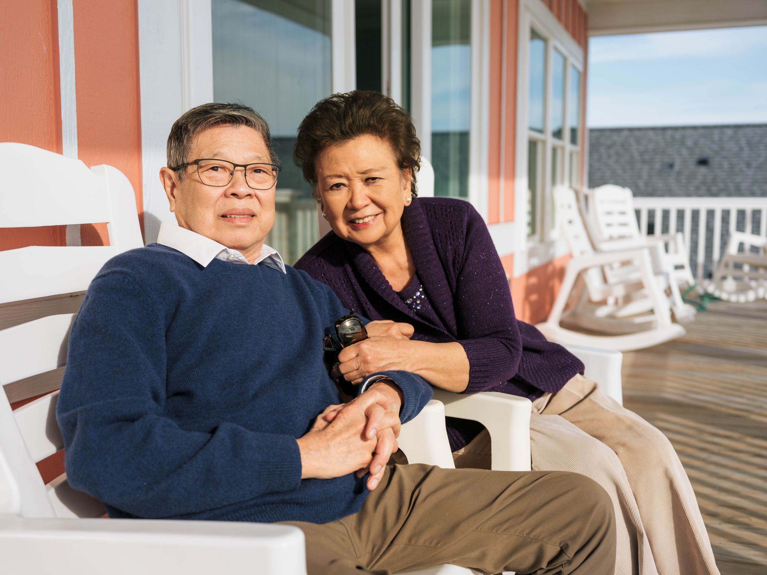 Elderly Asian couple smiling and holding hands on a porch with white rocking chairs on a sunny day.