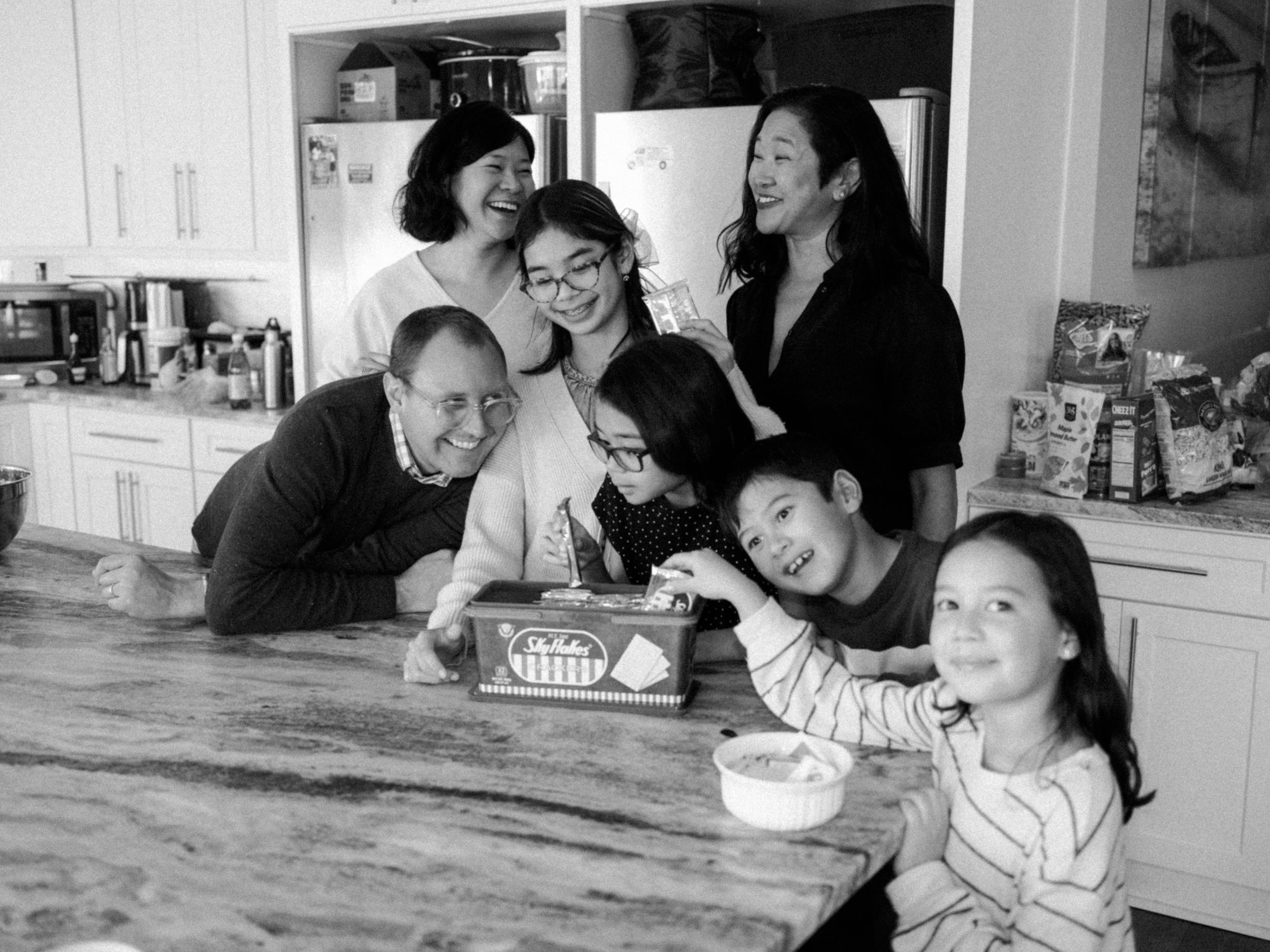 Black and white photo of a multigenerational Asian family gathered around a kitchen counter, smiling and sharing a joyful moment.