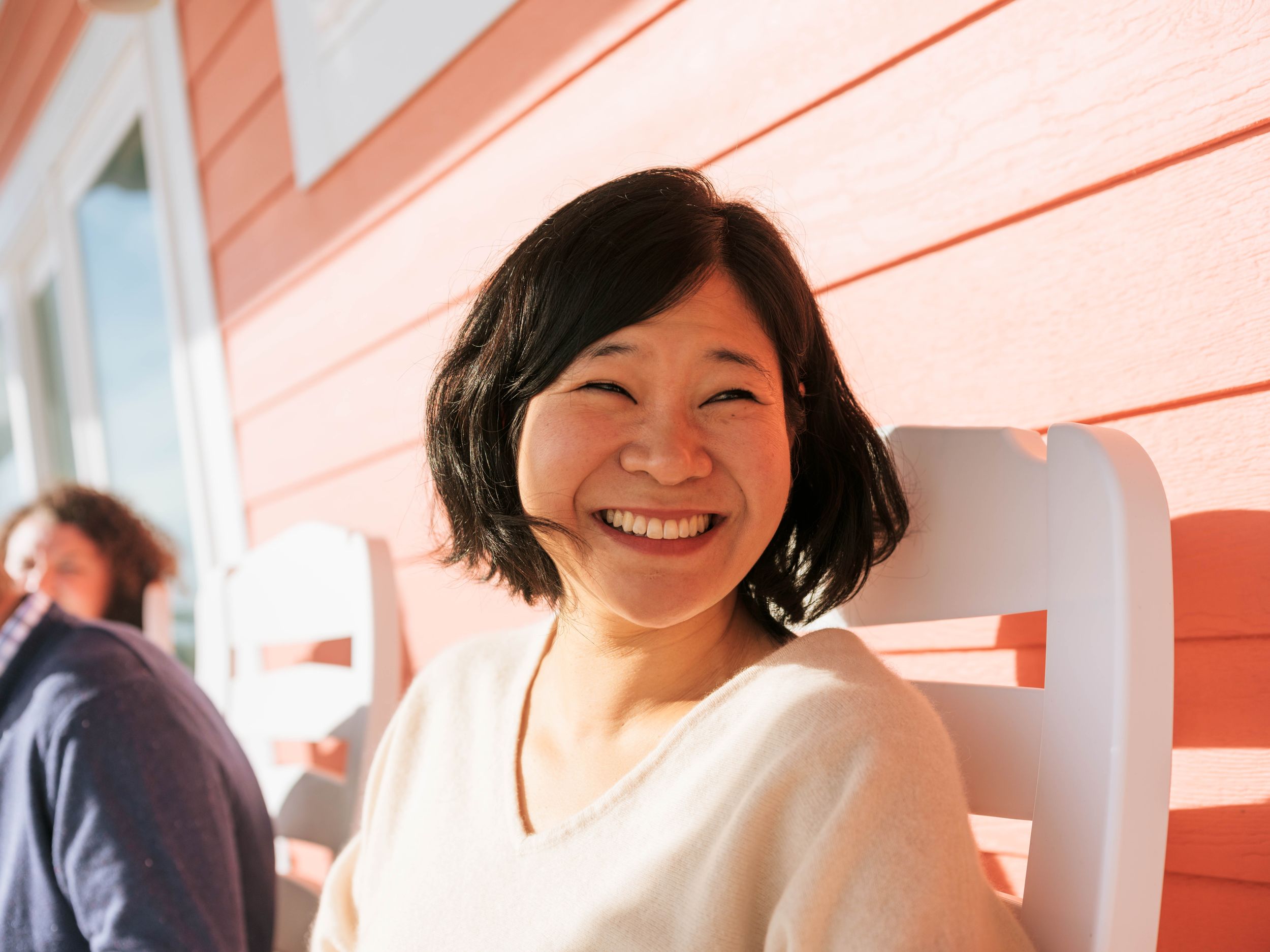 Smiling young Asian woman with short black hair sitting in a white chair outdoors near a red building.