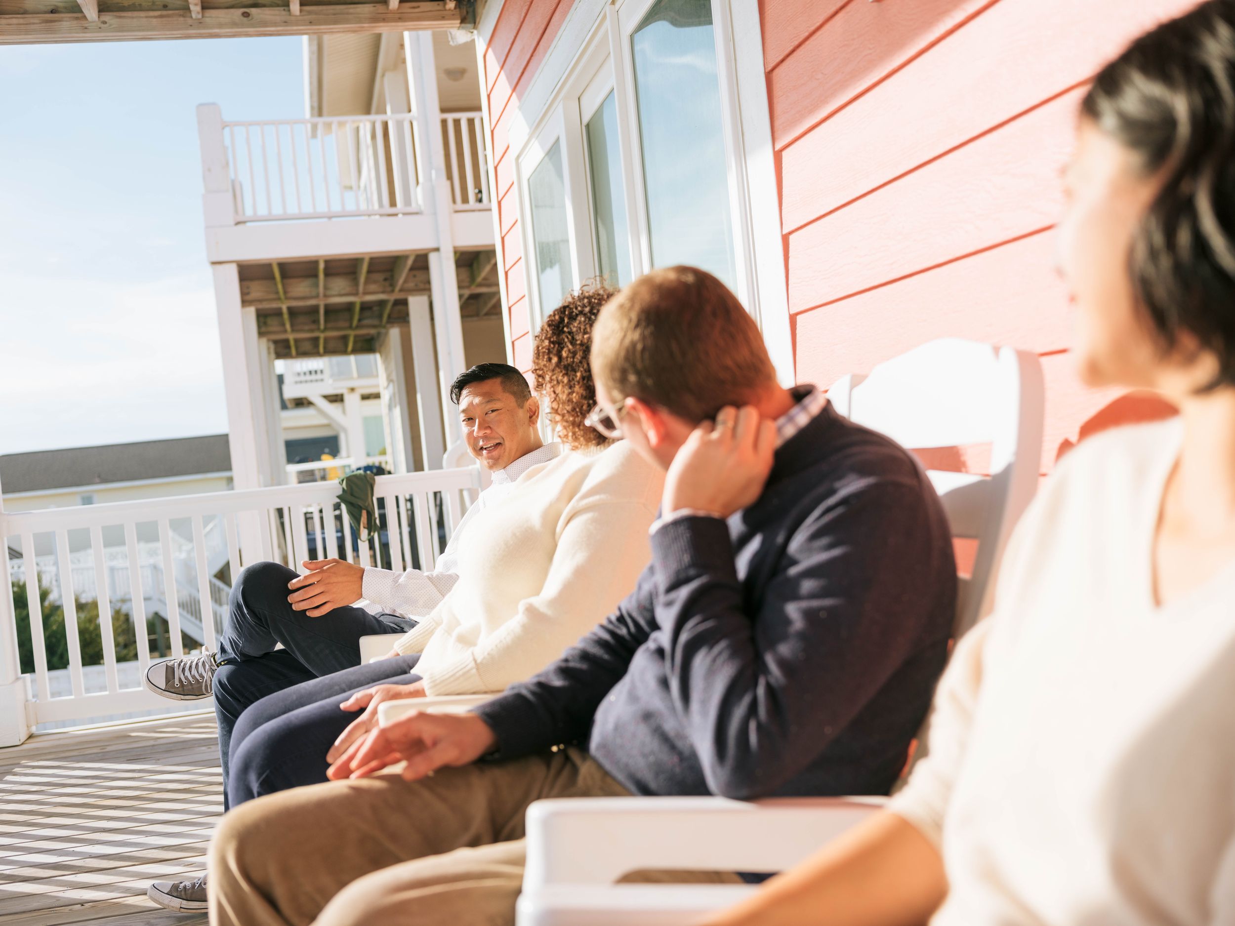 Group of friends relaxing on a sunny porch of a red beach house, enjoying conversation outdoors.