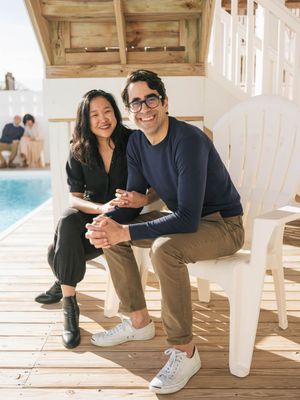 Two people smiling and sitting together on a wooden deck beside a pool on a sunny day.