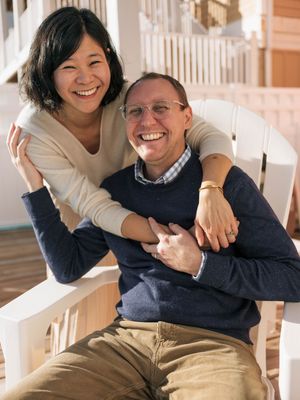 Happy couple smiling on a porch, woman hugging man from behind as he sits in a white chair outdoors.
