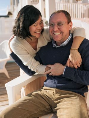 A happy couple embracing and laughing together on a sunny outdoor porch, wearing casual sweaters and khaki pants.