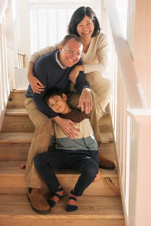 Happy mixed-race family of three posing together on wooden staircase at home, smiling warmly.