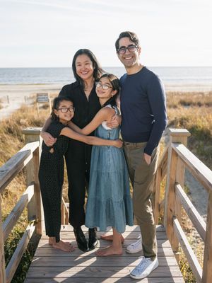 Family of four smiling together on a beach boardwalk on a sunny day, with ocean and sand dunes in the background.