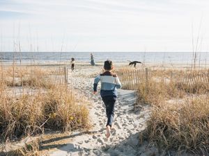 Child runs barefoot down sandy beach path through dunes toward ocean on a sunny day.