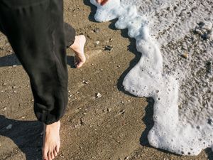 Barefoot person in black outfit walking on sandy beach with ocean foam waves washing ashore.
