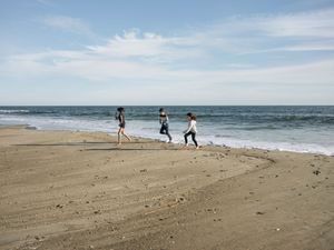 Three people walking along a sandy beach shoreline with gentle ocean waves and a partly cloudy sky.