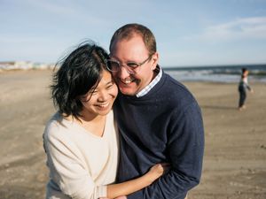 Happy couple embracing and laughing together on a sunny beach, with ocean waves visible in the background.