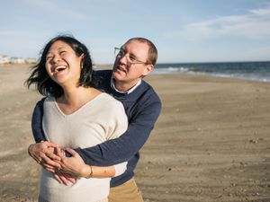 Couple laughing and embracing on a sandy beach with ocean waves in the background on a sunny day.