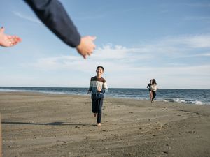 Child running barefoot on a sandy beach toward outstretched arms with another person in the background.
