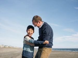 A man and young boy laugh together on a sandy beach under a bright blue sky.