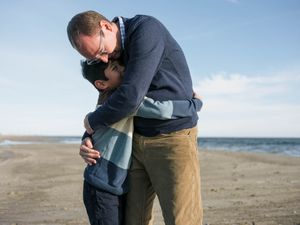 A man and woman share a warm embrace on a sandy beach under a clear blue sky.