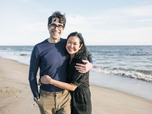 Couple embracing and smiling on a sandy beach with ocean waves in the background on a sunny day.