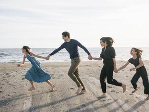 Group of friends holding hands and running barefoot on a sandy beach near the ocean on a sunny day.